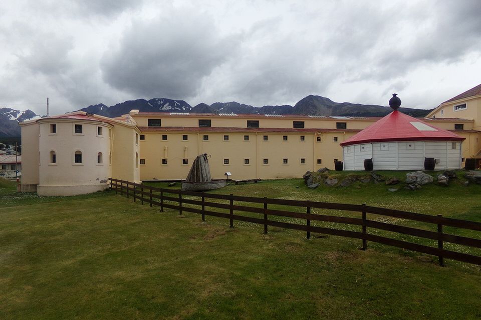 A long cream-coloured building with rows of small windows and a red conical roof, a lawn and wooden fence in front, snowy mountains behind under heavy cloud