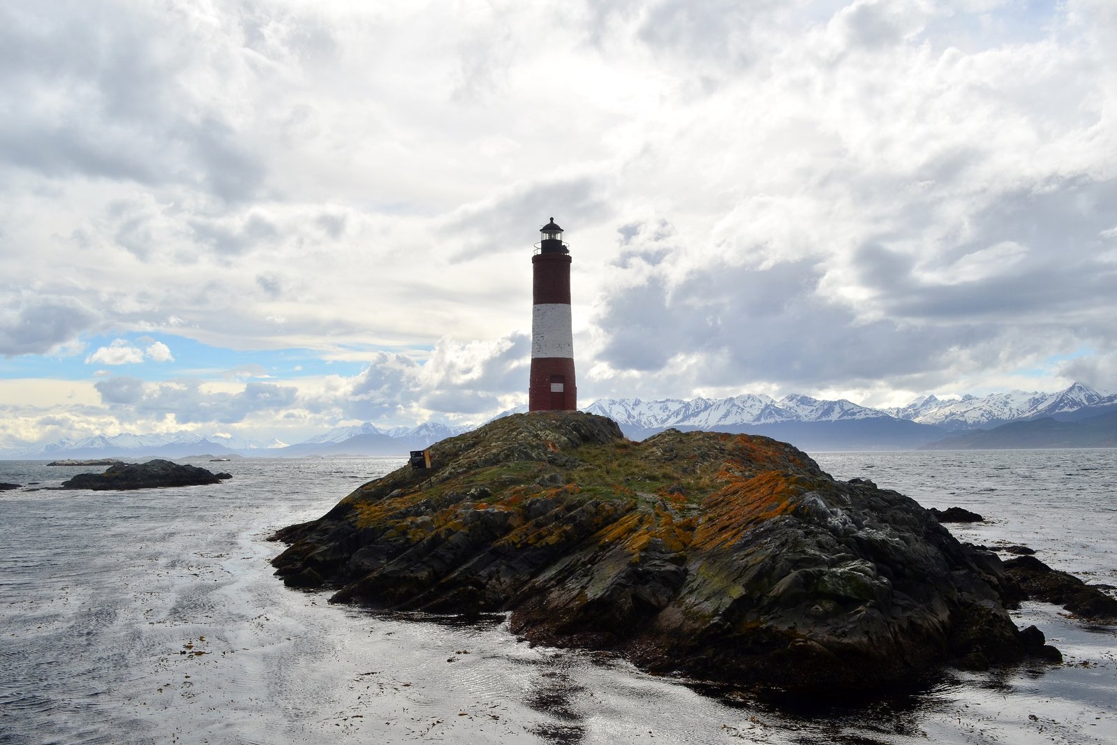 A red and white striped lighthouse on a low lichen-orange rock in grey water, a line of snow-capped mountains behind and a broken cloudy sky above