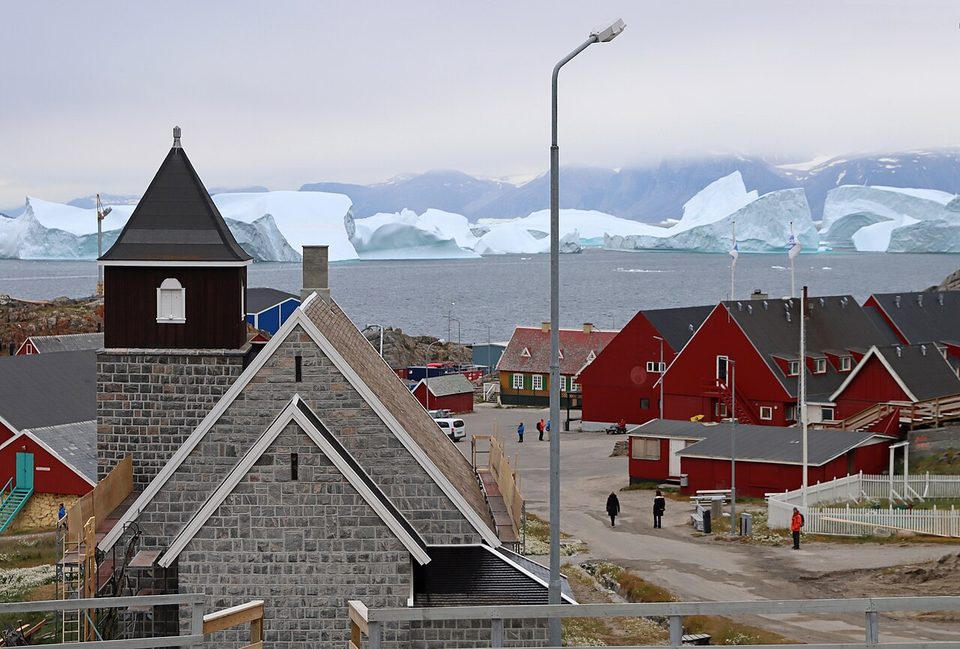 The town's church and red houses on rock above the water with large icebergs beyond