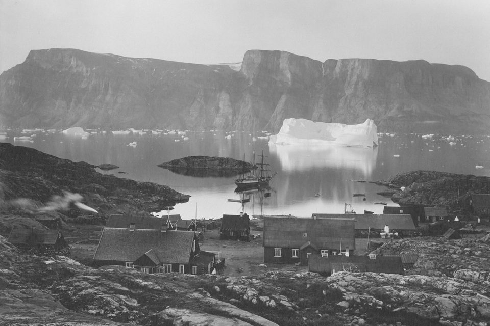 A black-and-white view of turf-roofed houses, a three-masted ship at anchor and a tabular iceberg below cliffs across the fjord