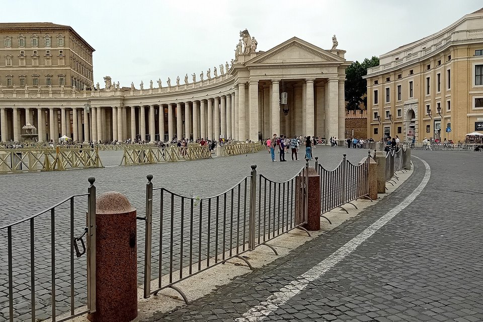 A pale stripe curving across grey cobbles beside low metal railings, the end of a curved colonnade and a tall building behind under a grey sky