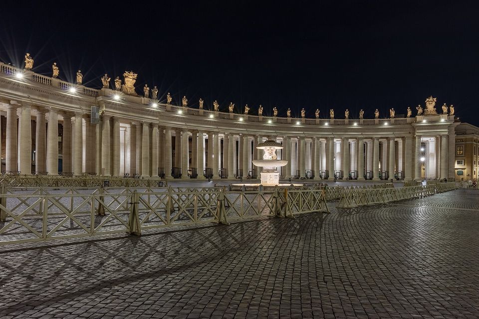 A curved colonnade of tall columns topped with statues, lit from below against a black sky, with a fountain and crowd barriers in the foreground