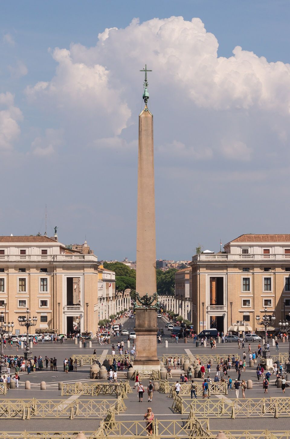 A tall pale stone obelisk topped with a cross against blue sky and a white cloud, the ends of the colonnade at either side and a few small figures at its foot