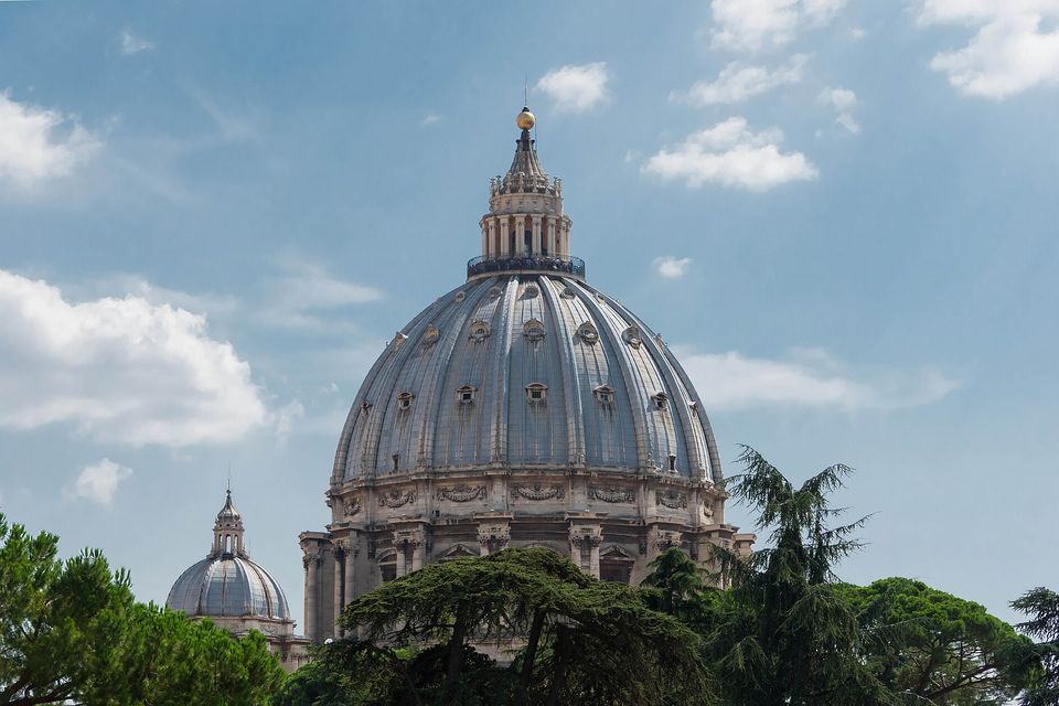The grey ribbed dome of a large basilica topped by a lantern and cross, framed by dark cedar branches against a blue sky with scattered white cloud