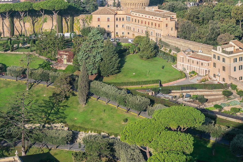 A hillside of green lawns, low clipped hedges and umbrella pines in front of pale ochre palace buildings and a long stone wall