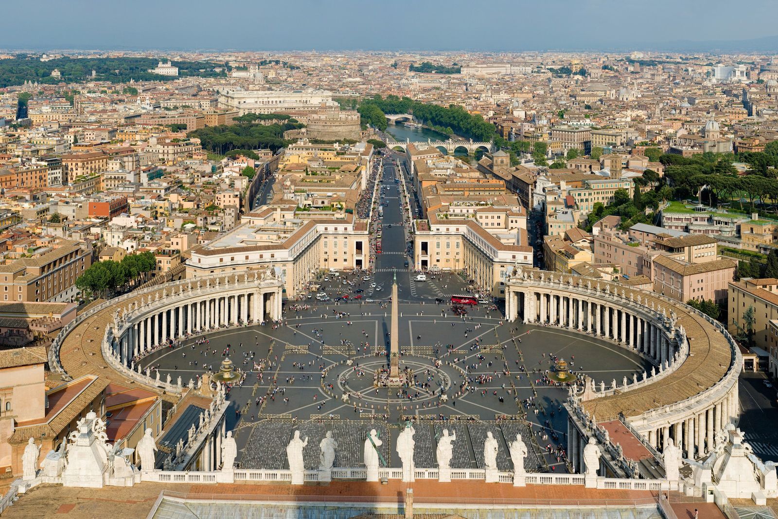 Aerial view of a large oval square ringed by curved colonnades, an obelisk at its centre, with a long avenue running away between the crowded rooftops of Rome