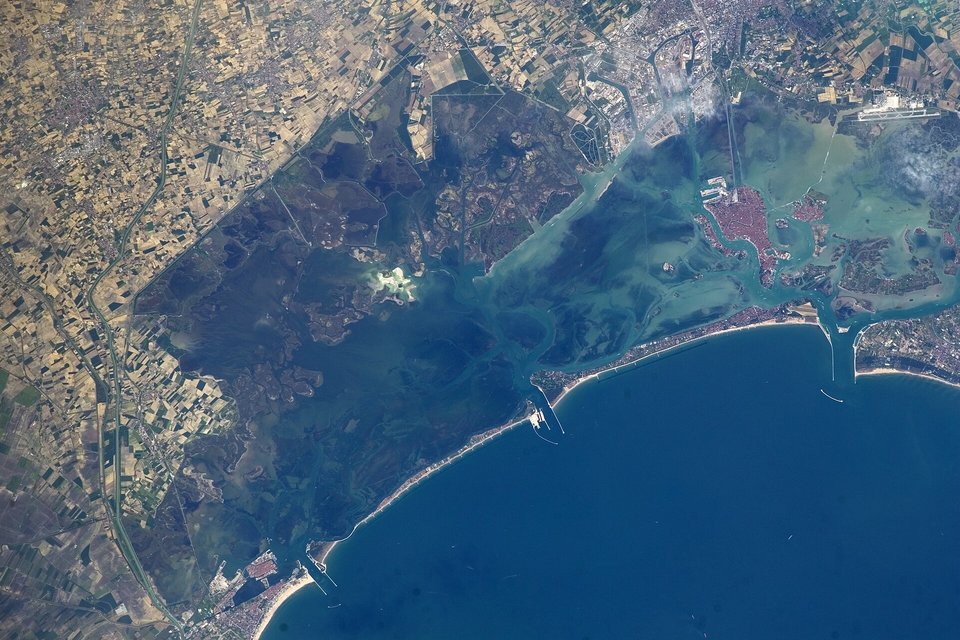 A view from orbit of a shallow blue-green lagoon with the pinkish patch of Venice at upper right, a long thin barrier island and the deep blue Adriatic beyond, farmland on the mainland at left