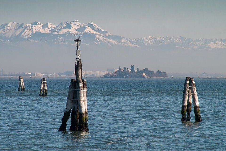 Three bundled dark wooden posts, one carrying a lamp, and two more clusters standing in calm blue water, a low island and snow-covered mountains behind in pale haze