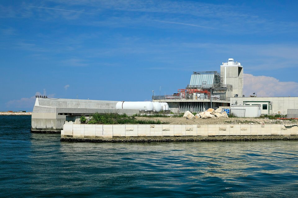 Low pale concrete works and a tower building on the shore of a lagoon inlet, calm green water in the foreground under a blue sky