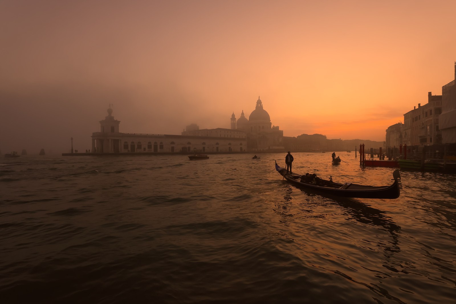The Punta della Dogana and the dome of Santa Maria della Salute in orange dawn fog across dark rippled water, a gondolier standing in a gondola in silhouette on the right