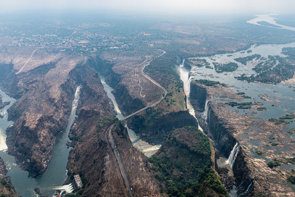 An aerial view of a zigzag series of narrow rust-brown gorges cutting away from the falls, a road and an arched bridge crossing them, hazy plateau beyond