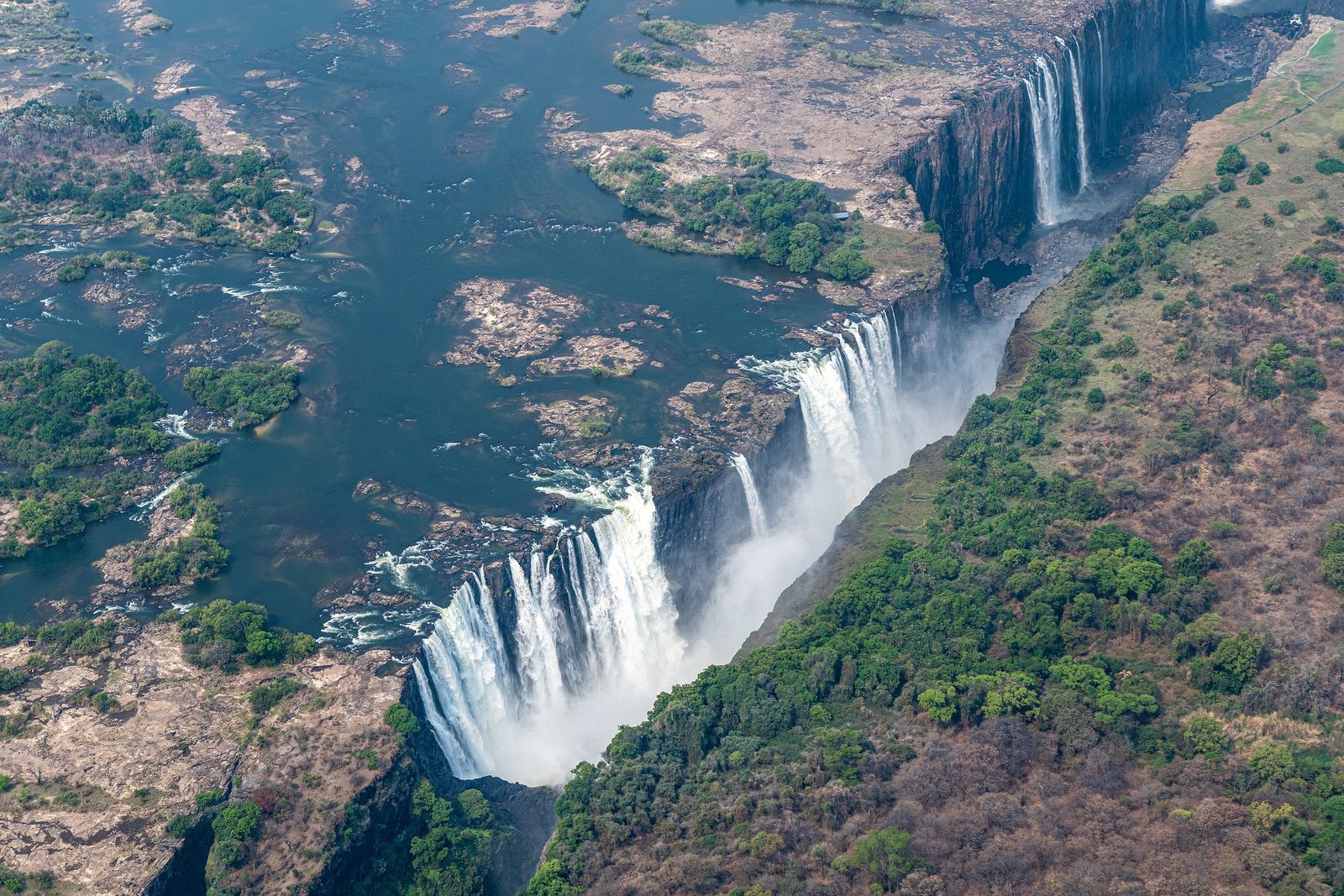 An aerial view of Victoria Falls: a broad curtain of white water dropping into a narrow chasm, dry-season islands and green forest on either side of the river