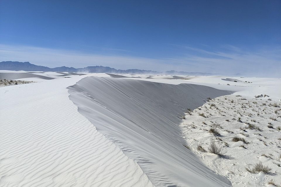 A smooth grey slip face curves away beside rippled white sand, tufts of grass on a flat at right and mountains on the horizon with sand blowing off distant dunes