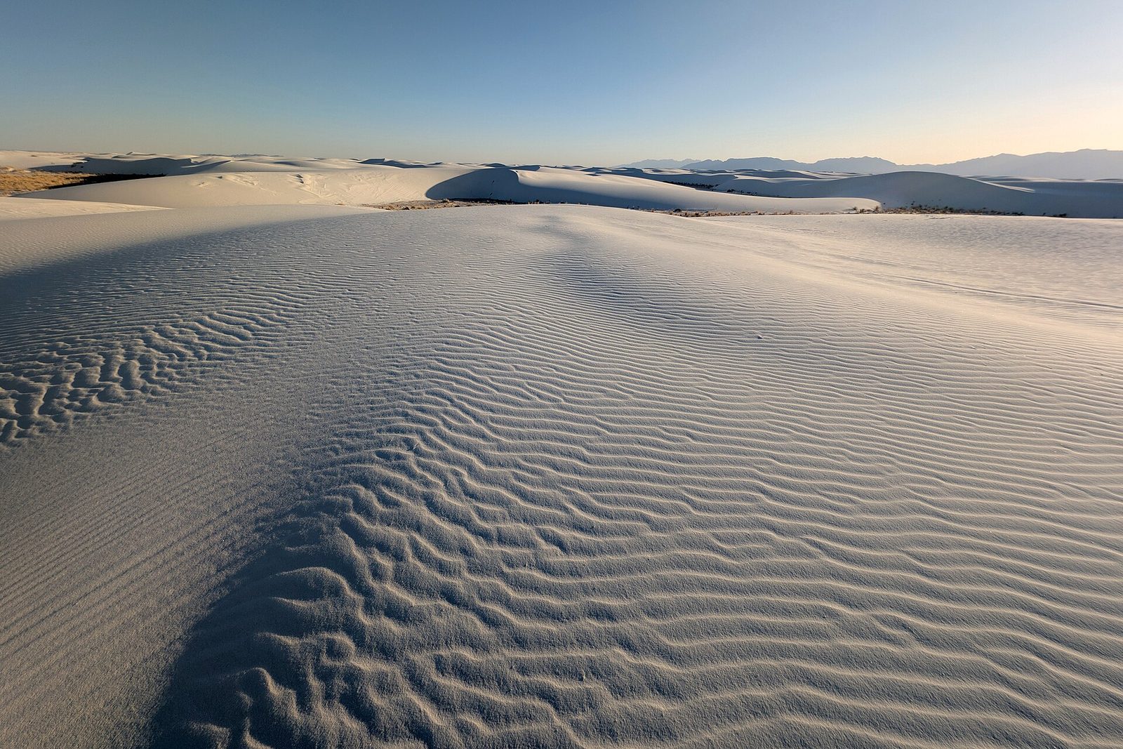 Low evening sun rakes across rippled white gypsum sand in the foreground, the dunefield rolling away in white waves to blue mountains on the horizon
