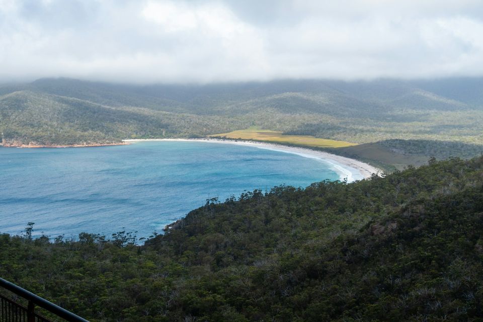A curved white beach and a blue bay seen from a lookout under low cloud, a platform rail in the corner