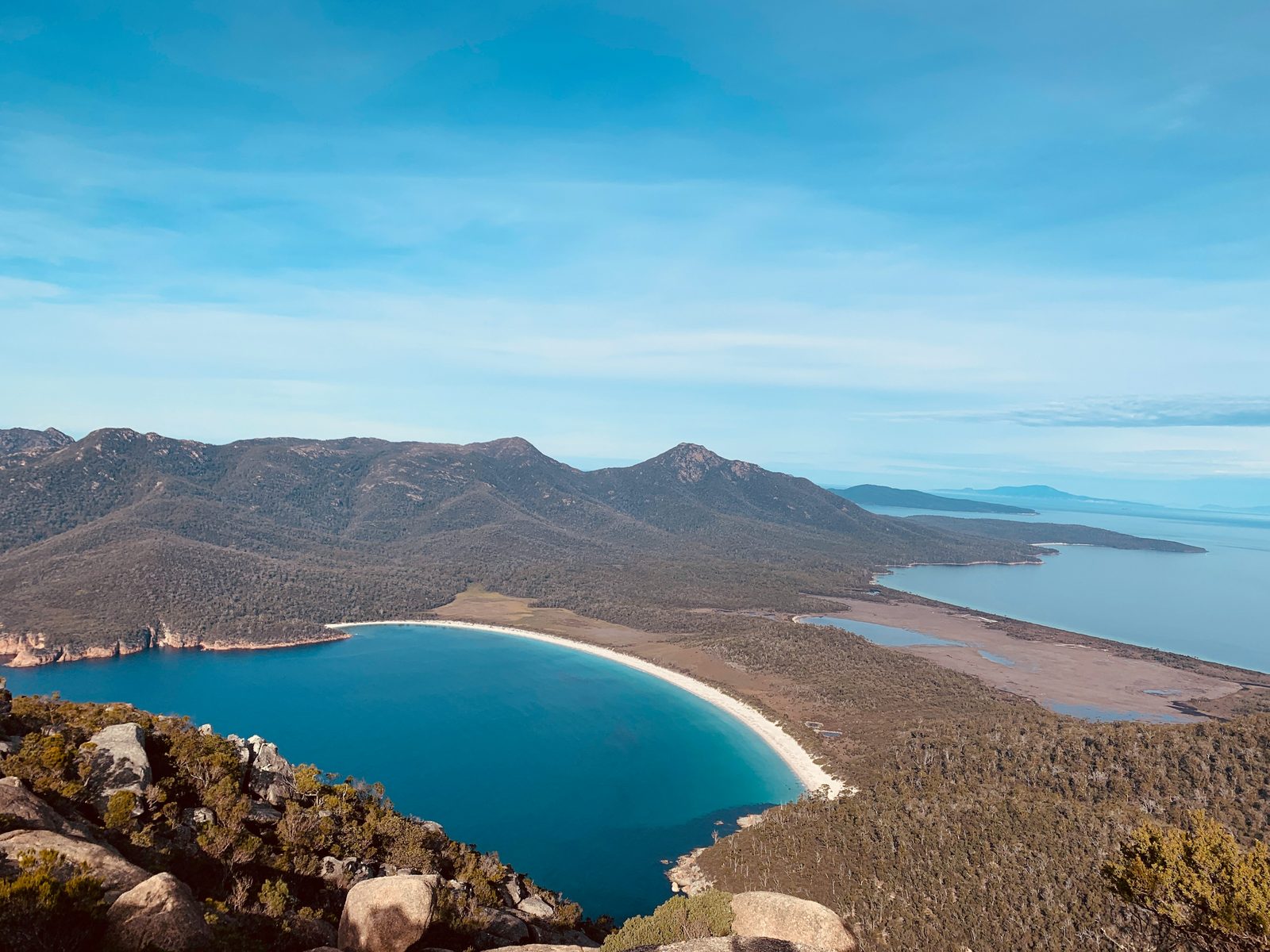 A curve of white sand around deep blue water below forested hills, granite boulders in the foreground