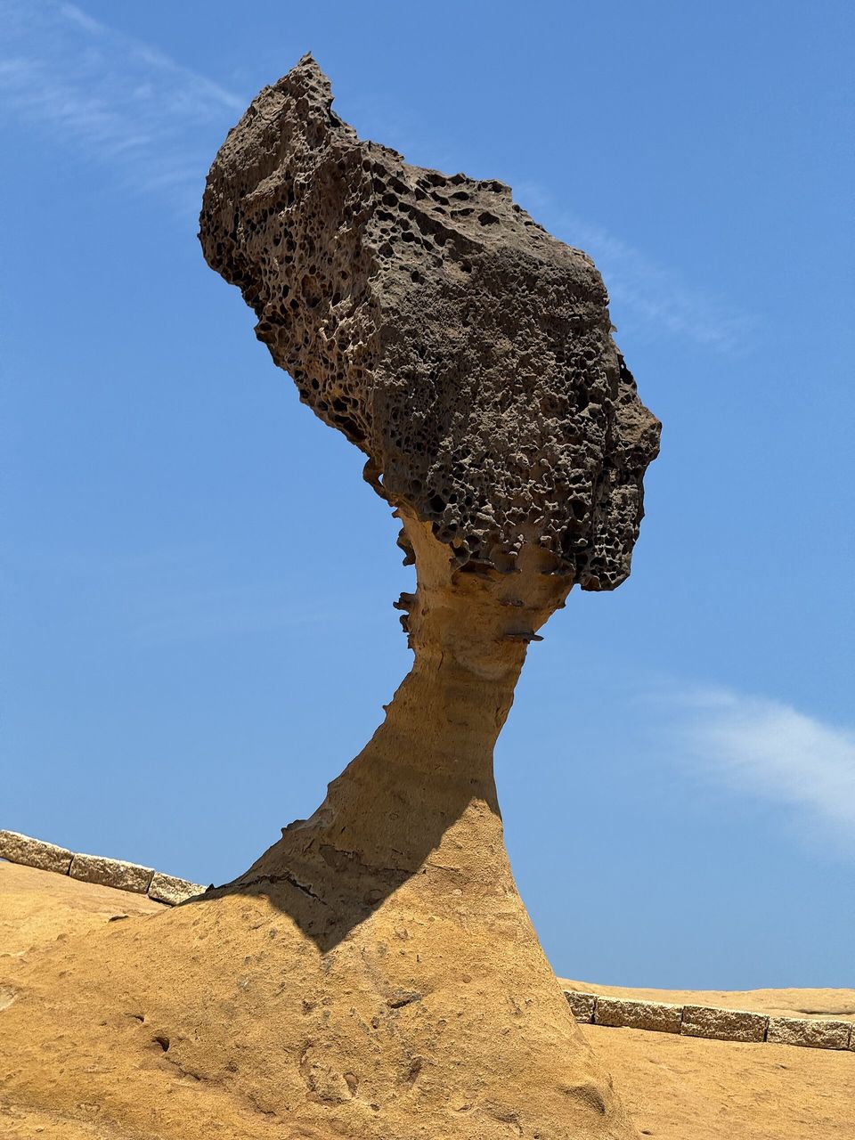 A tall rock shaped like a woman's head in profile, with a pitted dark cap on a slender pale sandstone neck, against a deep-blue sky, a low stone kerb around its base