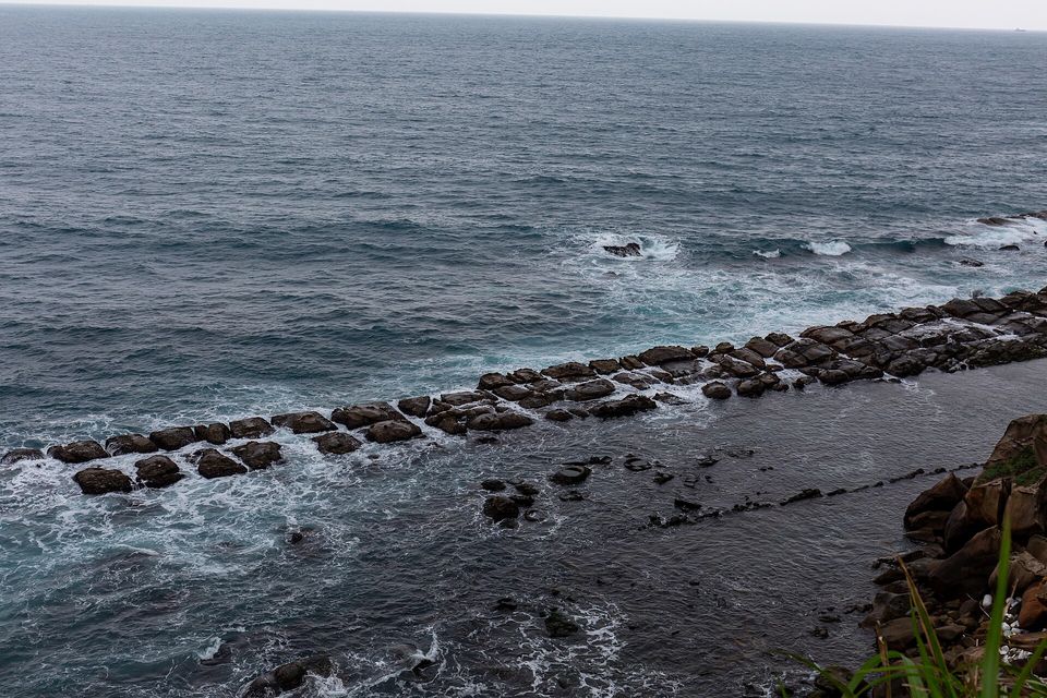 A tilted sandstone ridge with green algae at its base runs out into grey-blue sea, boulders in front and an island on the horizon