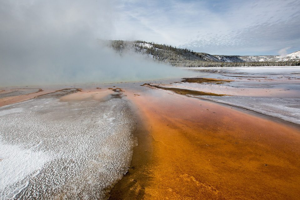 Bands of orange and rust-coloured microbial mat along a channel of steaming water edged with white mineral crust