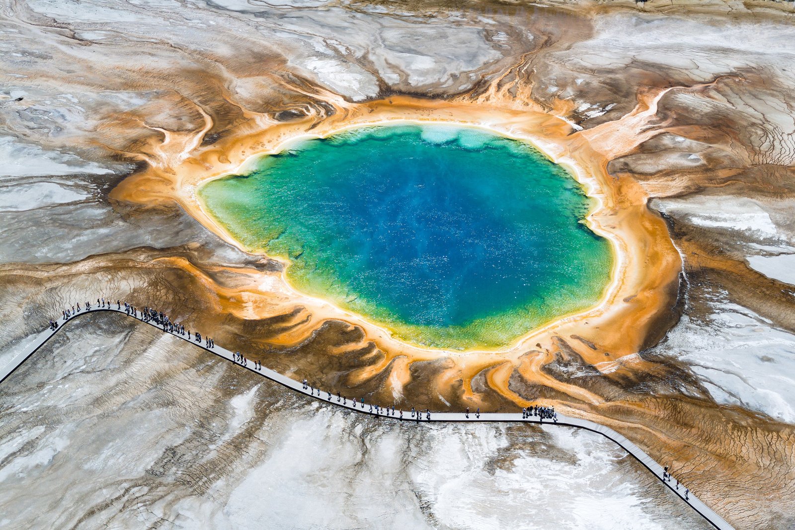 A blue and green hot spring ringed by orange and yellow microbial mats, with a boardwalk at the lower left and steaming ground around it