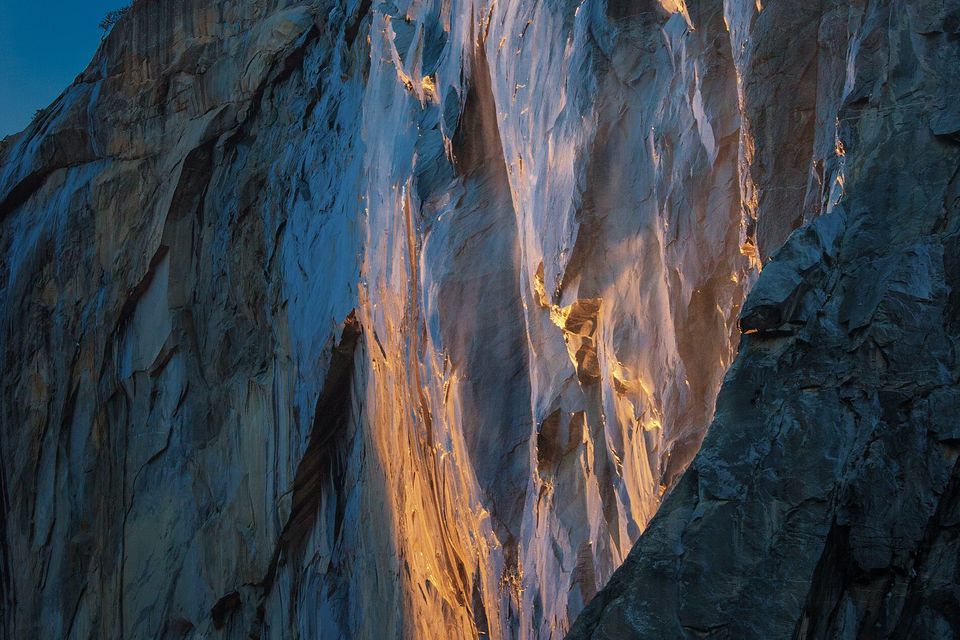 A steep granite wall streaked with bands of orange light, dark shadowed rock at the right and a strip of blue sky at the upper left