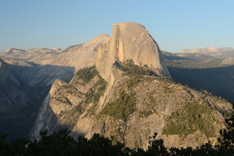 A rounded granite dome lit gold above a forested valley, more peaks and pale rock beyond under a clear sky, dark pine tops along the bottom edge