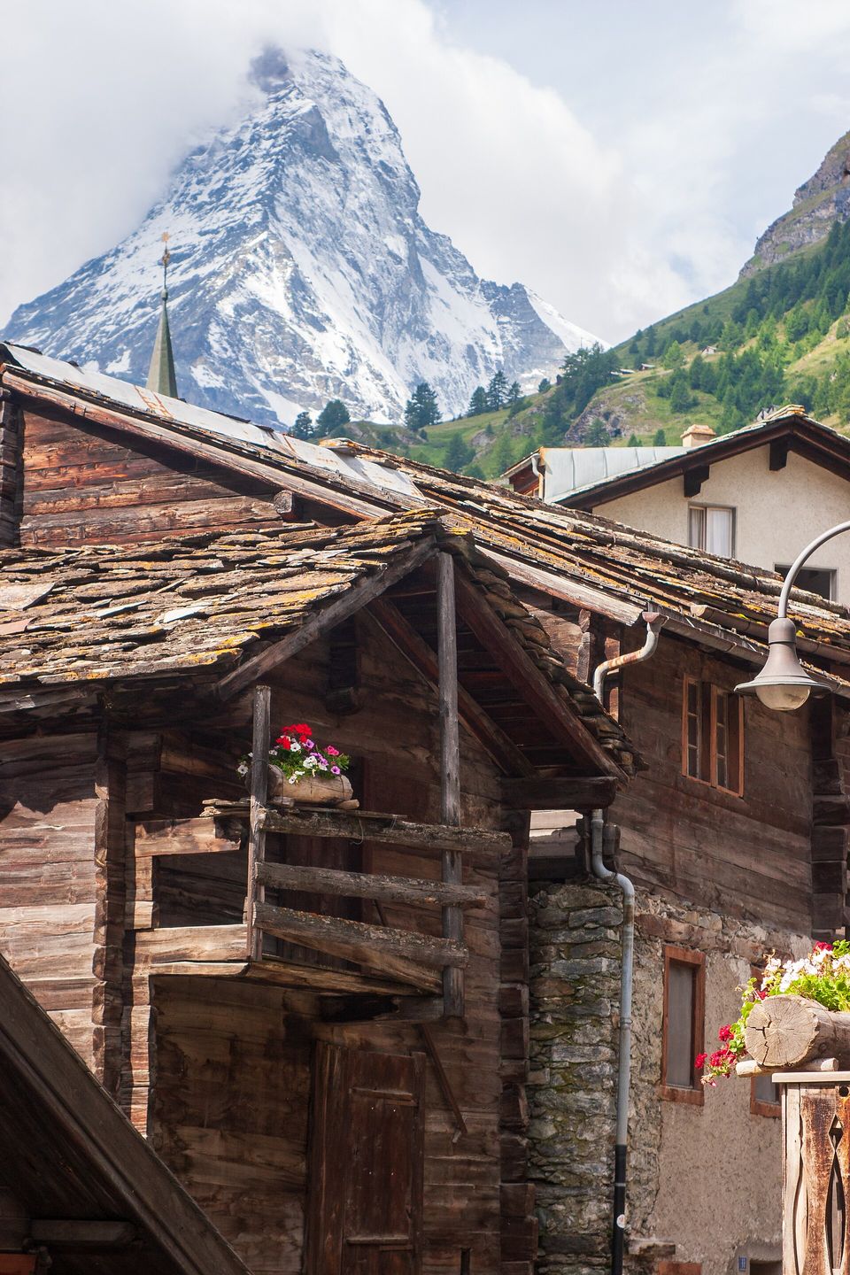 Dark timber houses with slate roofs and geraniums, a church spire, and the Matterhorn partly in cloud
