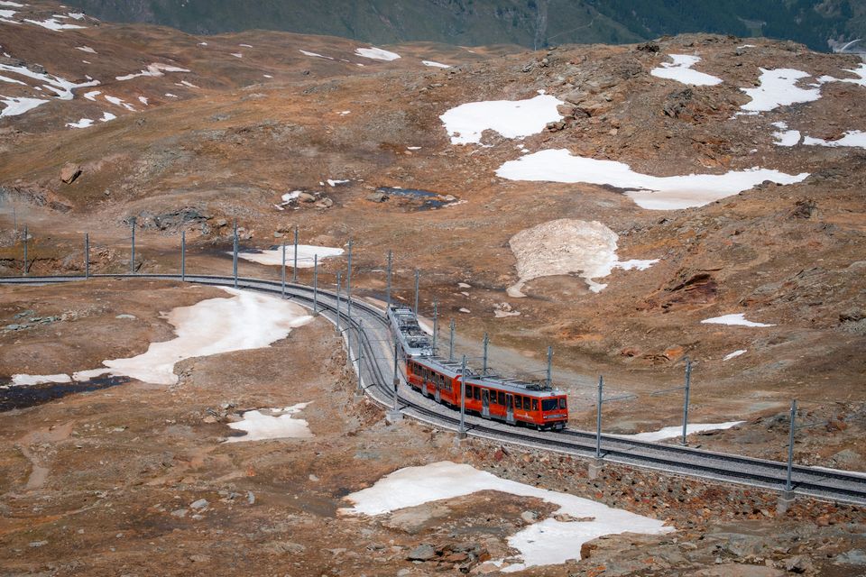 A red rack-railway train on a high ridge with patches of snow and peaks beyond