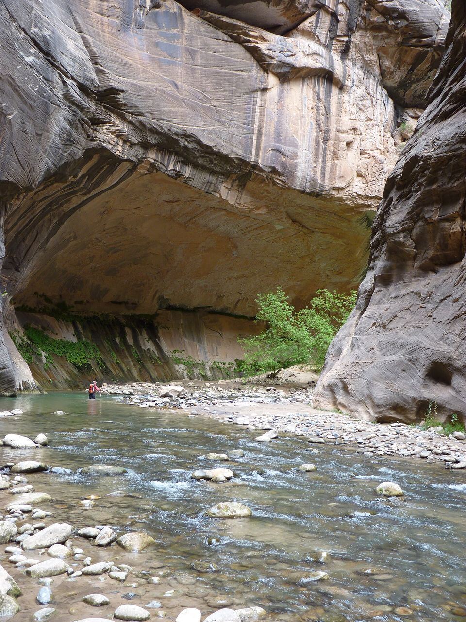 A shallow river over cobbles beneath a huge curved alcove in grey canyon walls, one small hiker far off in the water