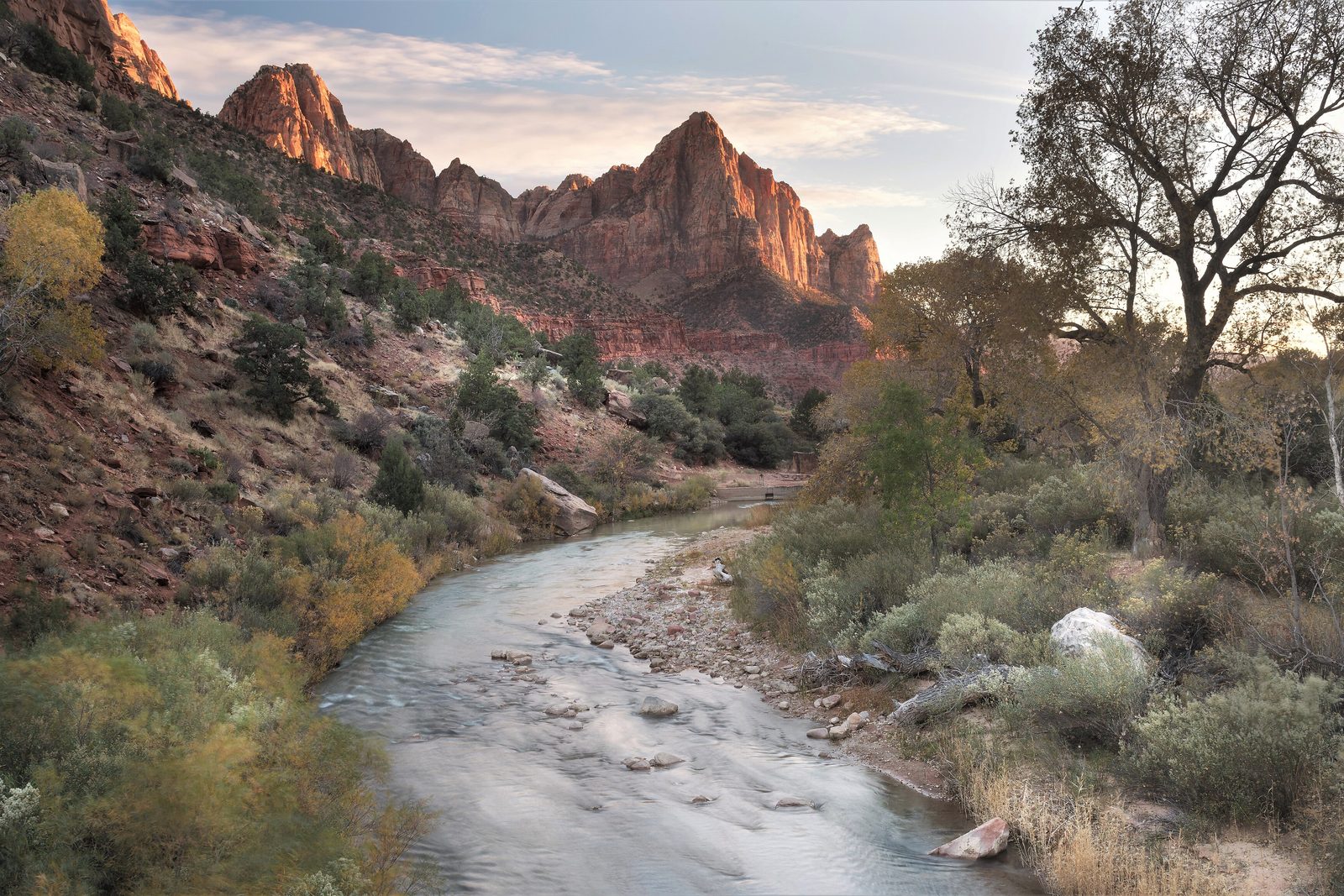 A red-rock peak lit at the top above a shallow river and golden cottonwood trees in soft evening light